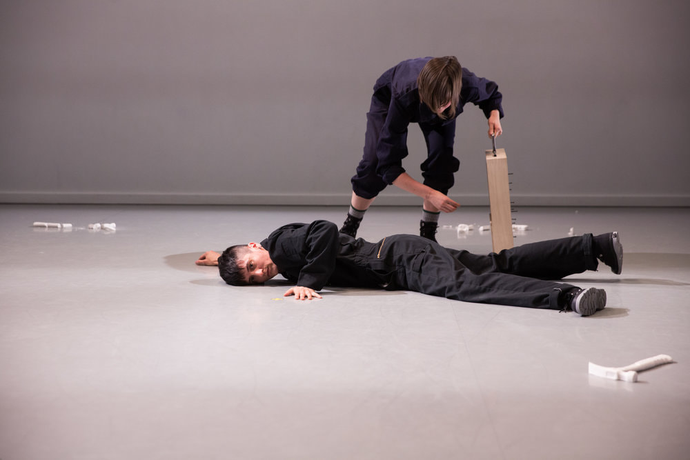 Amanda Hunt and IV Castellanos, Gibney Work Up, photo credit Scott Shaw. Two performers in jumpsuits interact during a performance. One is laying on the ground with eyes towards the viewer while the other stands overhead looking down.