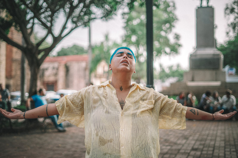 Agual Dulce in center of image with arms stretched out in a yellow button down shirt and blue hair. 