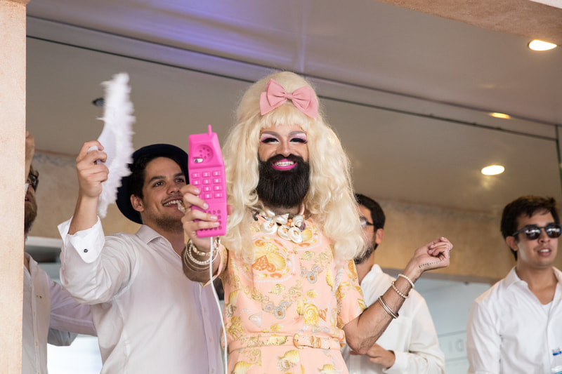 LINK to Sean Fader Artist Feature | A bearded Performer dressed in blond wig holds out their phone with a barbie pink phone cover.